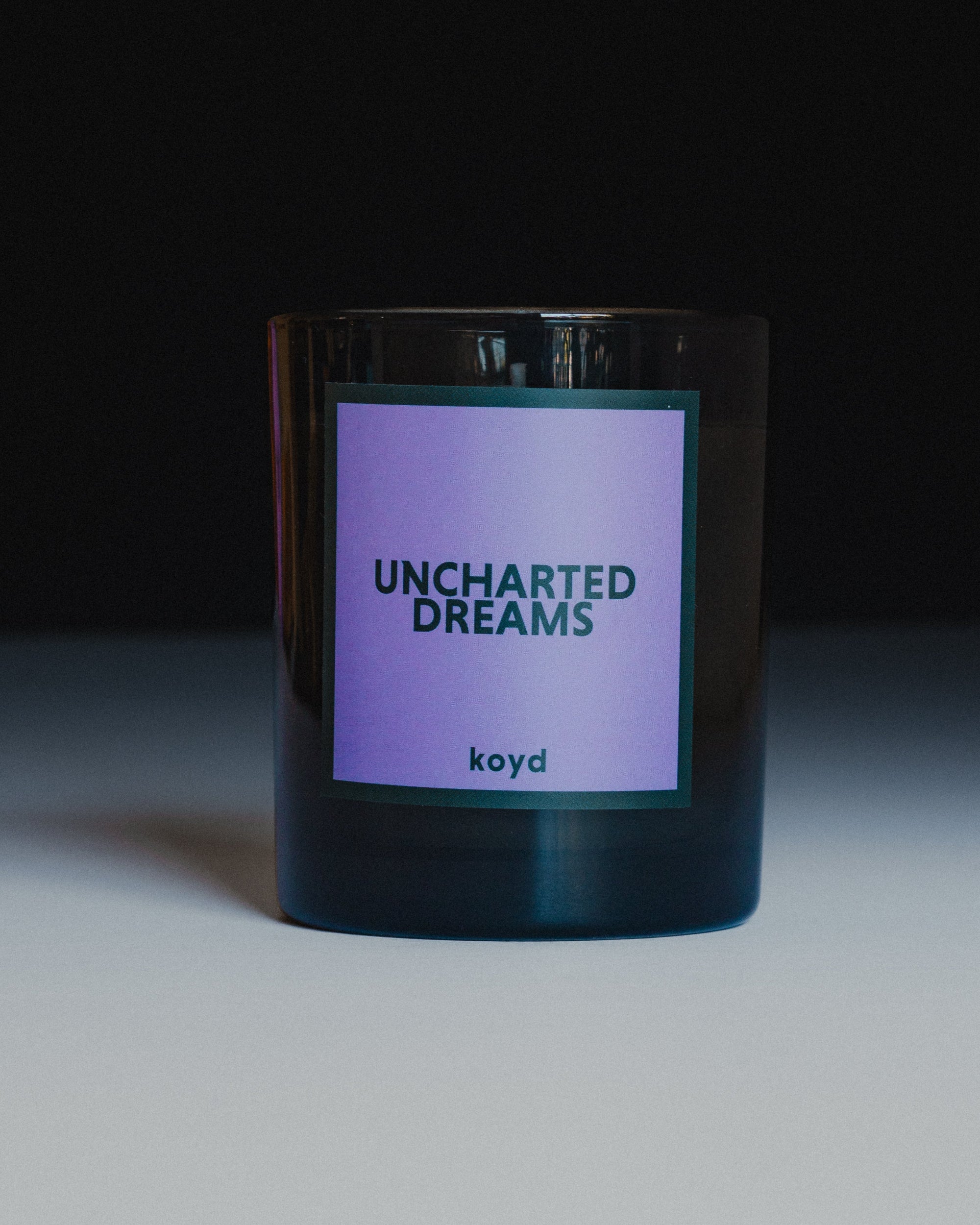 A dark glass jar candle with a lavender-purple label that reads 'UNCHARTED DREAMS' and 'koyd', photographed on a white surface against a dark background.