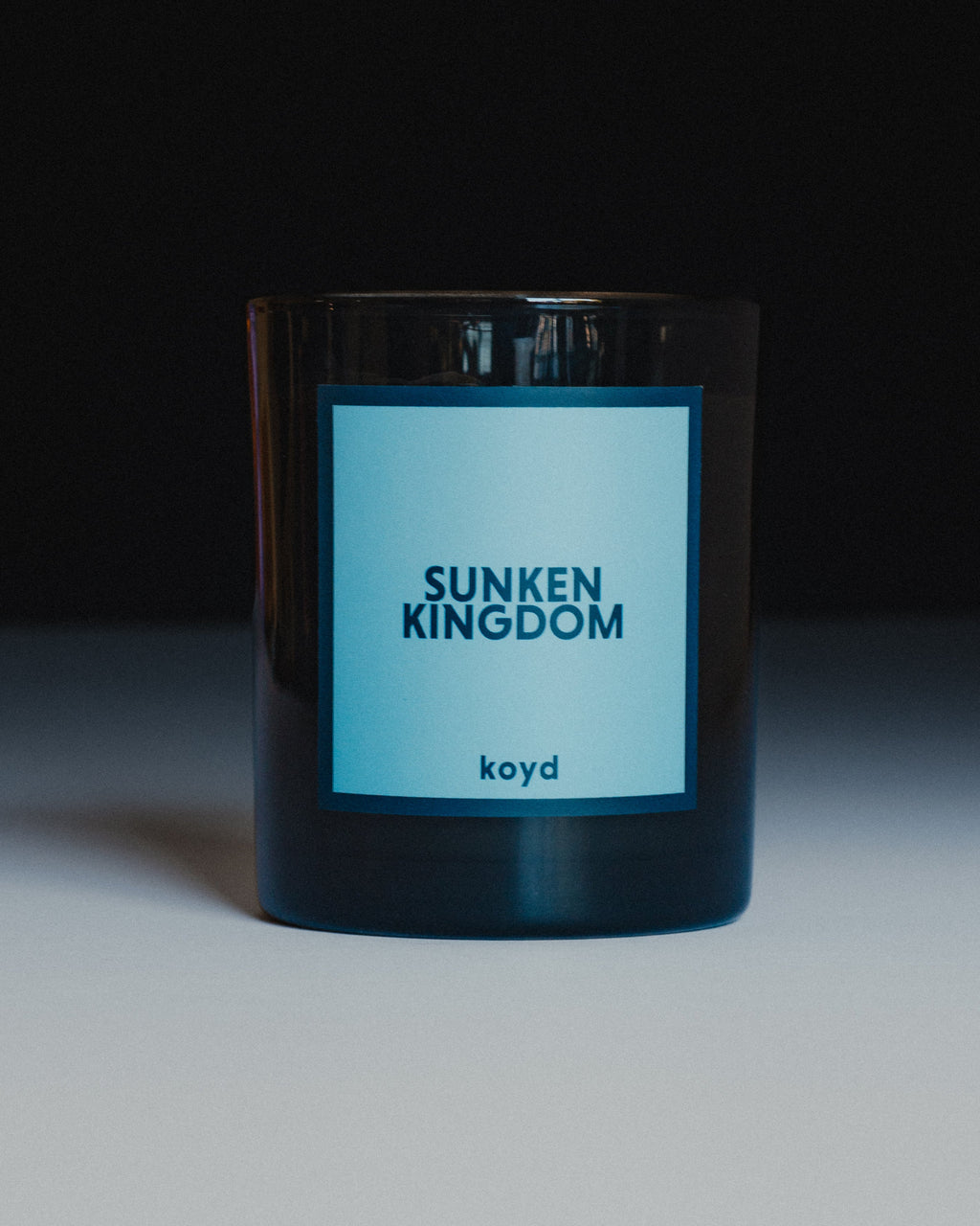 A dark glass jar candle with a light blue label reading 'SUNKEN KINGDOM' and 'koyd' below it, photographed against a dark background with a light surface below.