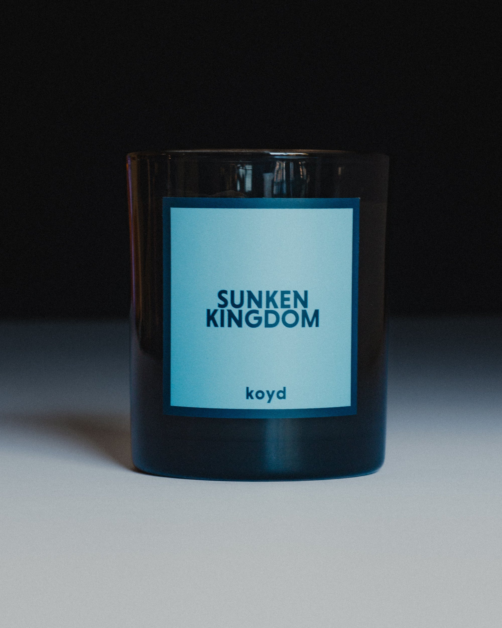 A dark glass jar candle with a light blue label reading 'SUNKEN KINGDOM' and 'koyd' below it, photographed against a dark background with a light surface below.