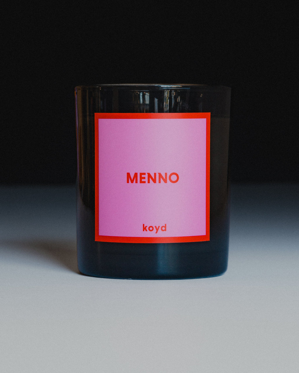A dark glass jar candle with a pink label featuring the text 'MENNO' and 'koyd' in red, set against a dark background with a light surface.