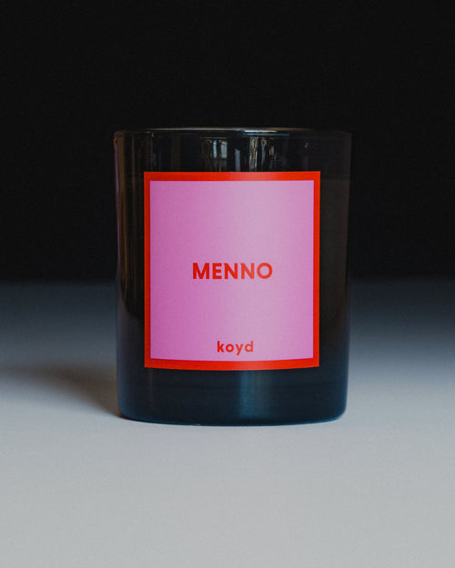 A dark glass jar candle with a pink label featuring the text 'MENNO' and 'koyd' in red, set against a dark background with a light surface.