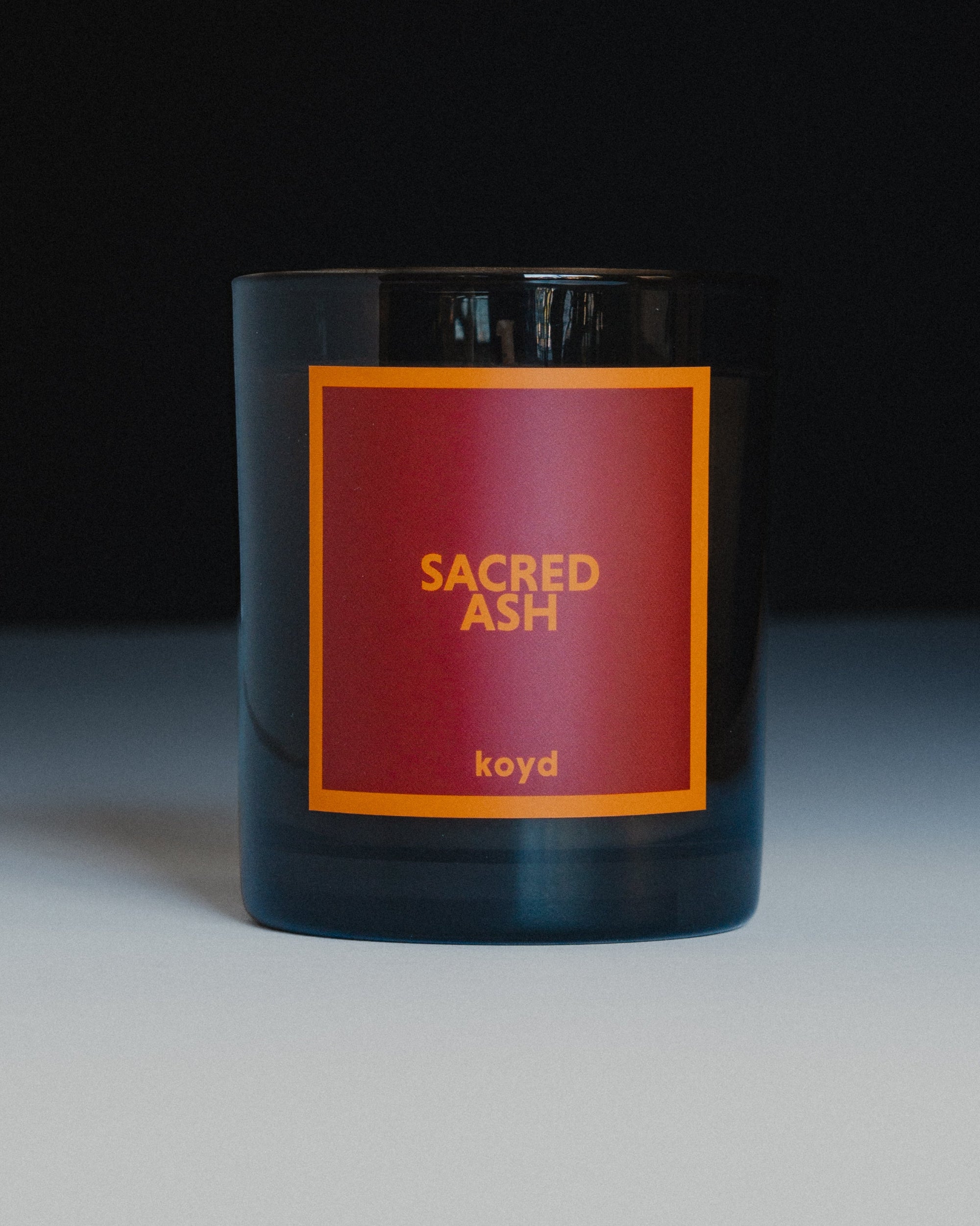 A dark glass candle jar with a square red label featuring the text 'SACRED ASH' and 'koyd' in orange, set against a dark background.