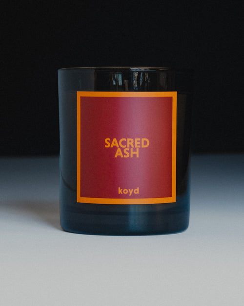 A dark glass candle jar with a square red label featuring the text 'SACRED ASH' and 'koyd' in orange, set against a dark background.