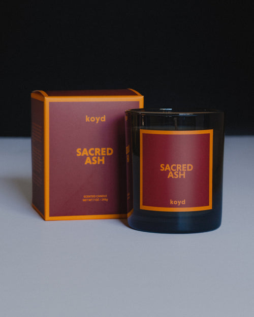 A black glass jar candle labeled 'SACRED ASH' by koyd, shown next to its matching dark red and golden yellow box on a  white surface against a black background.