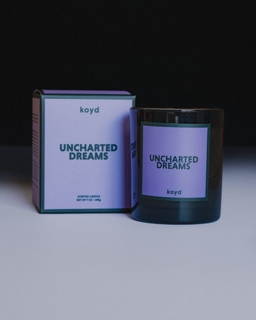 A dark glass jar candle and its matching product box from the brand koyd, featuring a soft purple label with the scent name 'UNCHARTED DREAMS' in dark teal text, set against a dark background.