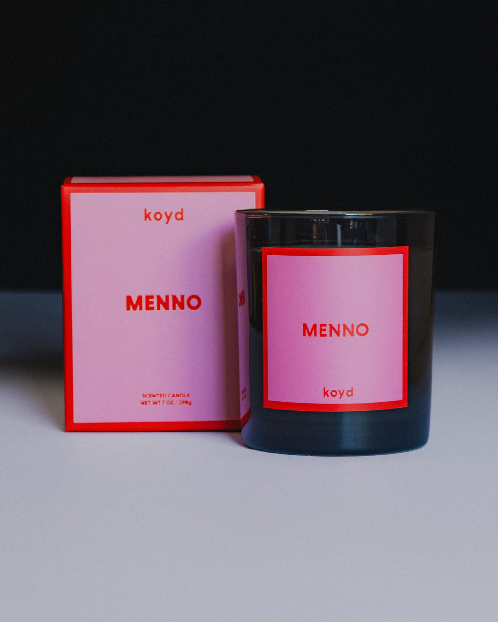 A black glass scented candle with a pink and red label reading 'koyd MENNO' stands next to its matching pink and red box on a white surface against a dark background.