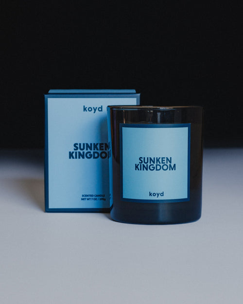 A candle in a dark glass jar with a light blue label reading 'SUNKEN KINGDOM' and 'koyd', shown next to its matching blue box on a white surface against a dark background.