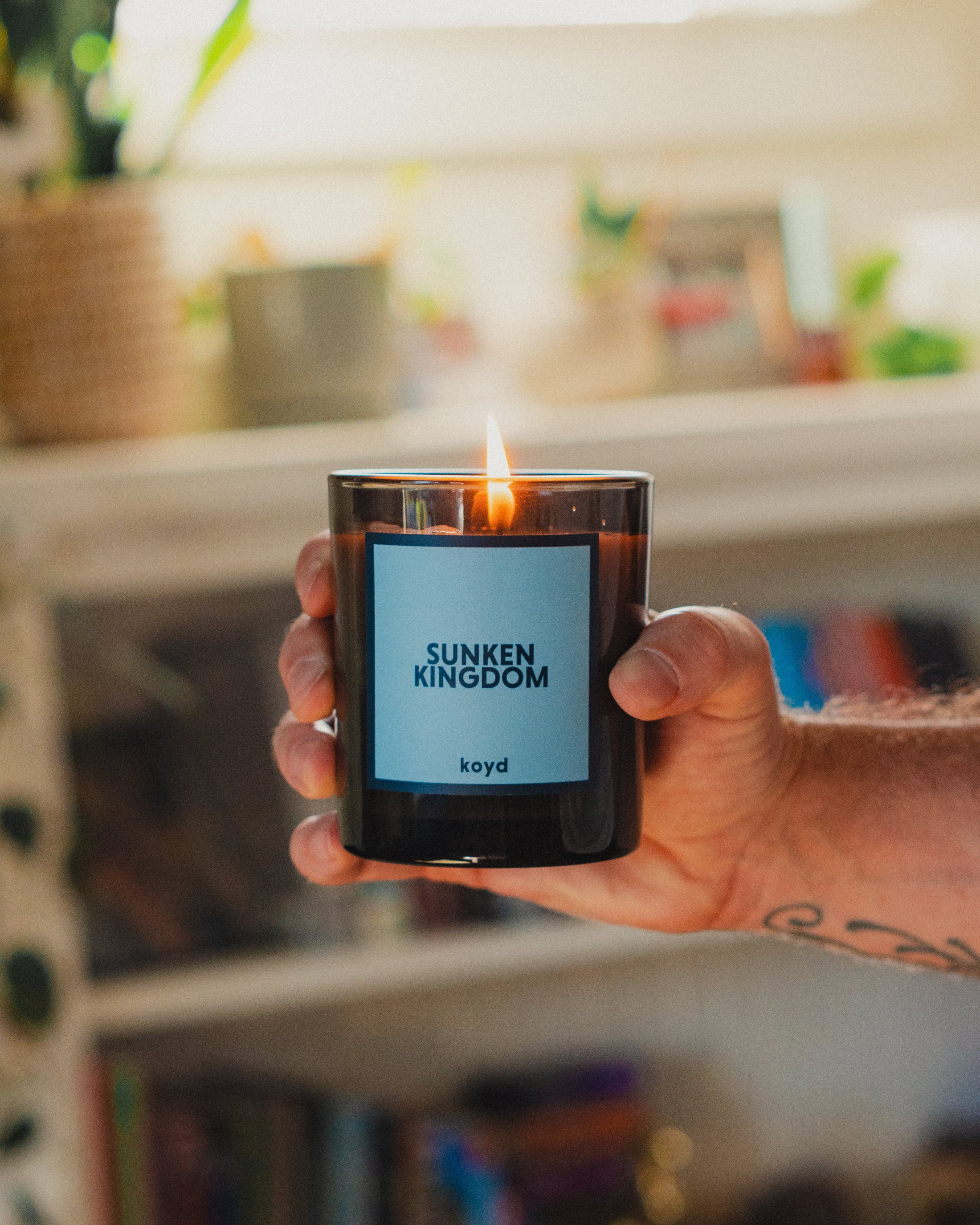 A hand holds a lit black glass candle jar with a light blue label reading 'SUNKEN KINGDOM' and 'koyd'. The background shows a softly blurred indoor shelf setting with books and plants.