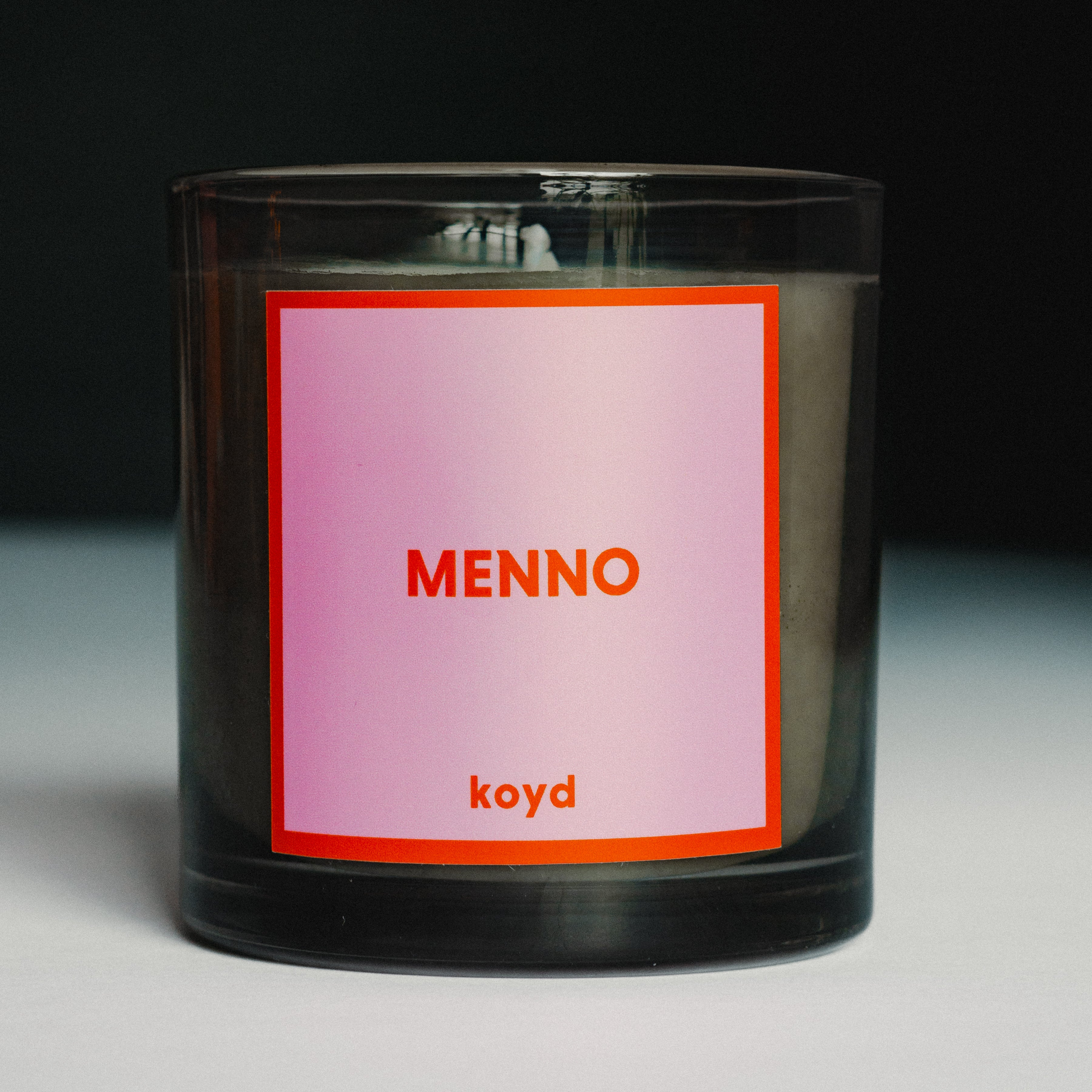 Black candle with a pink and red label featuring the candle name 'MENNO' and the brand name 'koyd' on a dark background