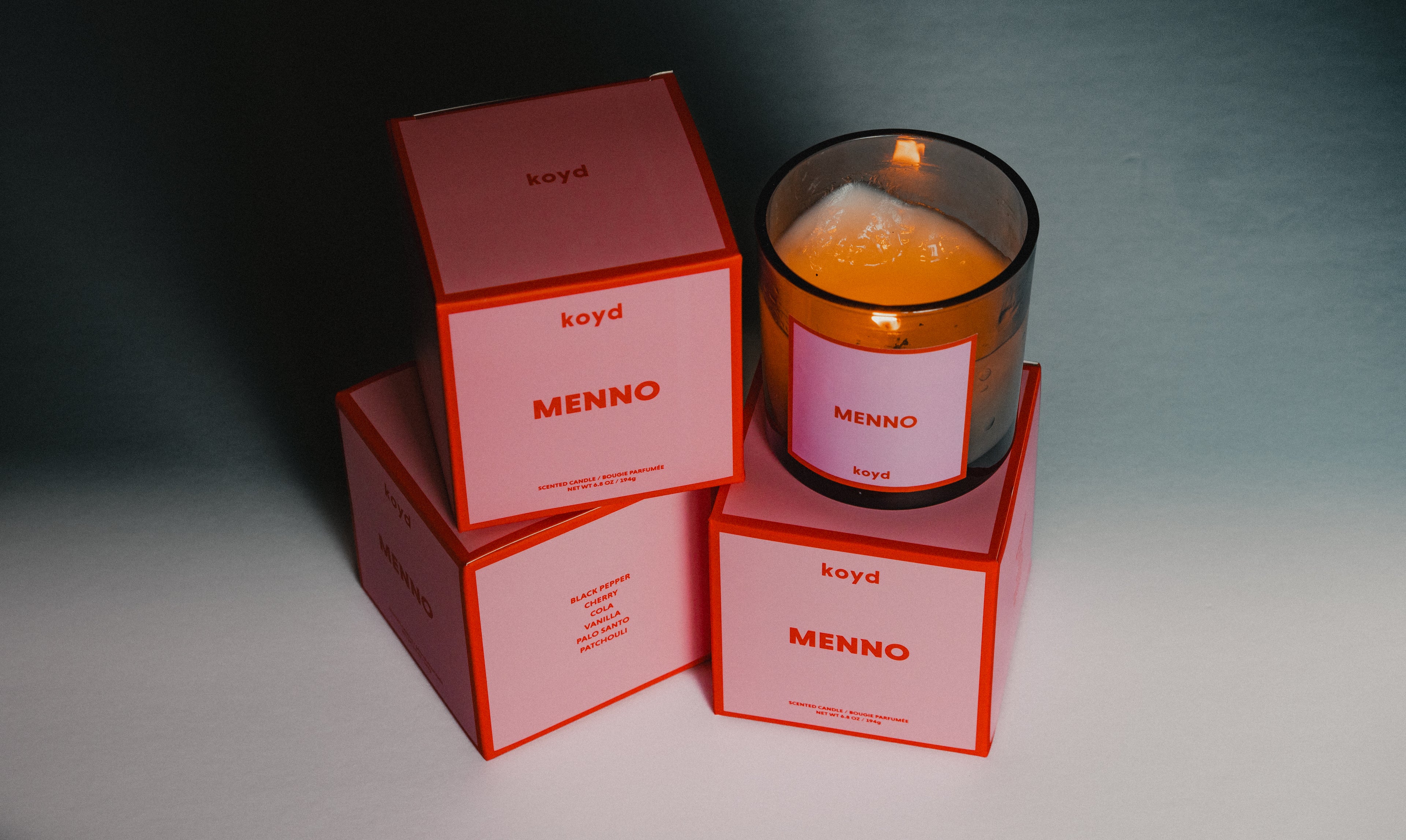 A black semi-transparent candle with pink and red labeling. The candle is lit and sitting on top of 3 boxes that also have pink and red labeling.