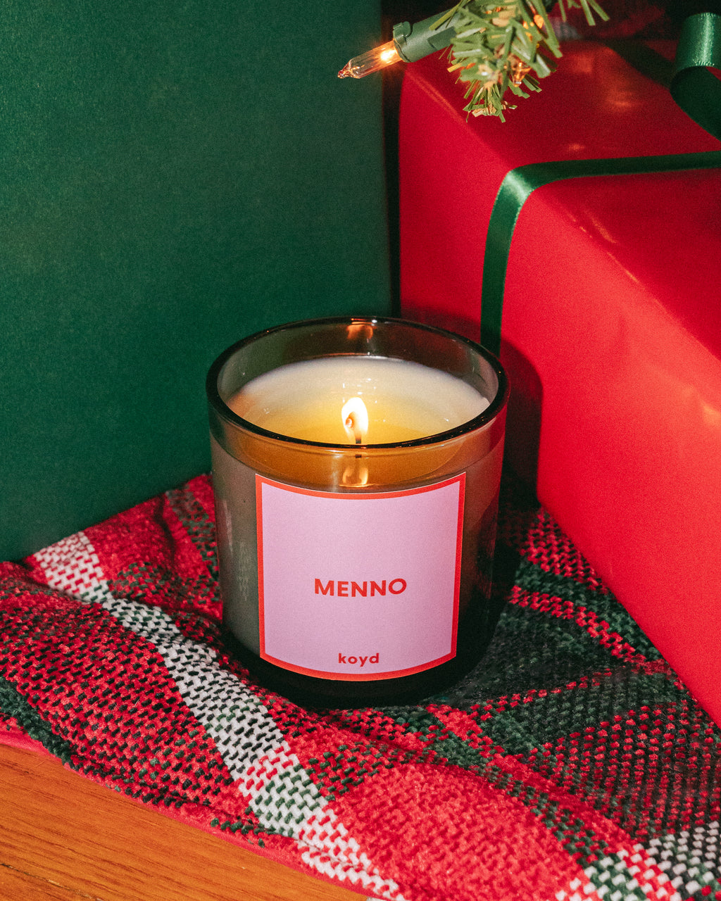 A black semi-translucent glass candle with pink and red labeling sitting on a plaid blanket next to gift boxes.