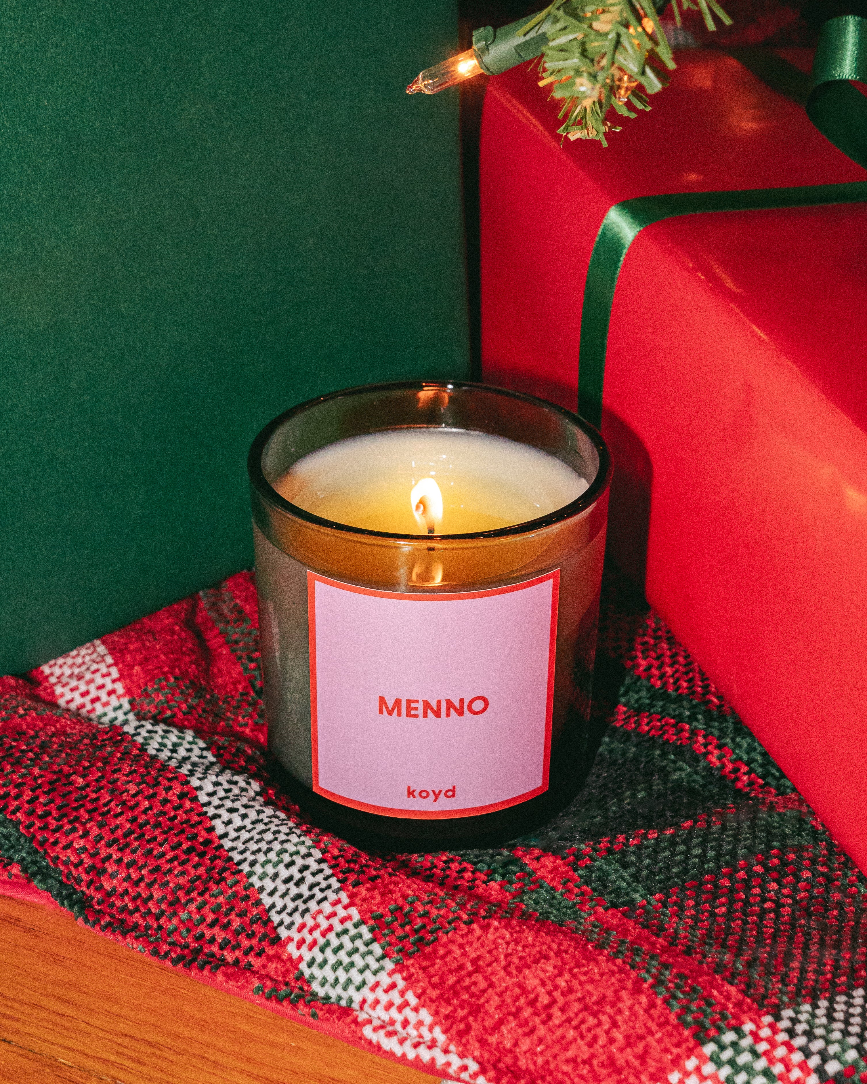 A black semi-translucent glass candle with pink and red labeling sitting on a plaid blanket next to gift boxes.